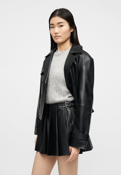 Jeune femme portant une veste en cuir noire sur un pull en maille gris et une jupe en cuir plissée noire avec ceinture, debout devant un fond uni.