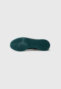 Sål af en mørk teal sneakers med strukturerede cirkulære grebmønstre og et Puma-logo i hælområdet mod en simpel hvid baggrund.