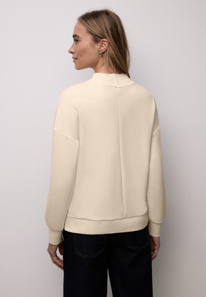 Lys cremefarvet pullover med høj hals, ribbet kant og ballonærmer. Blødt stof med en glat tekstur. Skræddersyet pasform, minimale syningsdetaljer.