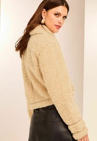 Beige faux fur jakke med høj krave, tekstureret stof og ribbede manchetter, parret med en sort lædernederdel.