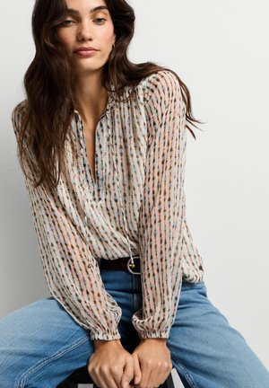 Kvinde med langt, mørkt hår iført en mønstret bluse med lange ærmer og blå jeans, der sidder med hænderne foldet foran en ensartet lys baggrund.