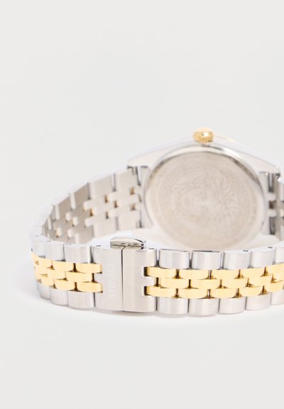 Μεταλλικό δίχρωμο λουράκι ρολογιού με ασημένιους εξωτερικούς κρίκους και χρυσούς κεντρικούς, με αγκράφα με χαραγμένο το όνομα της μάρκας.