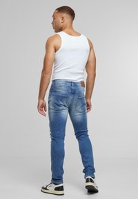 Slim fit kék farmernadrág, fakó mosással, öt zsebbel és bőr folttal a deréknál. Fehér ujjatlan pólóval és sportcipővel viselve.