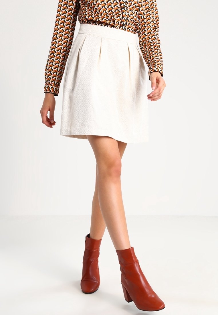 Kreemi pleatattu hame, jossa on teksturoitu pinta, yhdistettynä ruskeisiin nilkkureihin. Asusteessa on kuvioitu pitkähihainen paita.