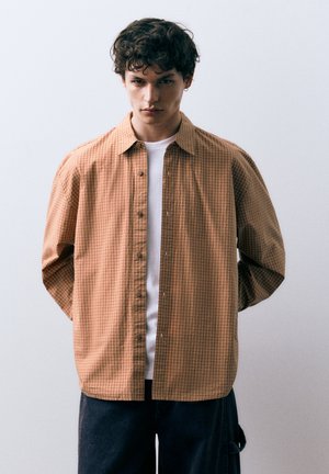Chemise marron à carreaux avec un devant boutonné, un col classique et des manches longues. Portée sur un T-shirt blanc. Design simple et décontracté.