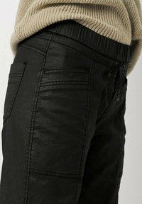 Schwarze tapered Hose mit elastischem Bund, ausgestattet mit einem Kordelzug, Seitentaschen und sichtbaren Nähten. Die Textur wirkt weich.