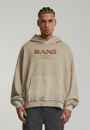 Beige oversized hoodie with a front pocket, featuring bold text "KANI" in brown. Soft fabric with a ribbed hem and cuffs.