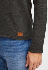 Donkergrijze langemouwen shirt van gebreide stof, met een gestikte zoom en een bruine leren label aan de onderkant. Gecombineerd met blauwe jeans.