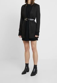 Blazer nero con un design su misura, dotato di una cintura, maniche lunghe e tasche. Indossato su un vestito nero strutturato e abbinato a stivali neri alla caviglia.