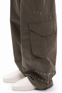 Široke cargo hlače tamno maslinasto zelene boje, s bočnim džepom i ukrasima u obliku prstenova na rubu. Glatka tekstura.