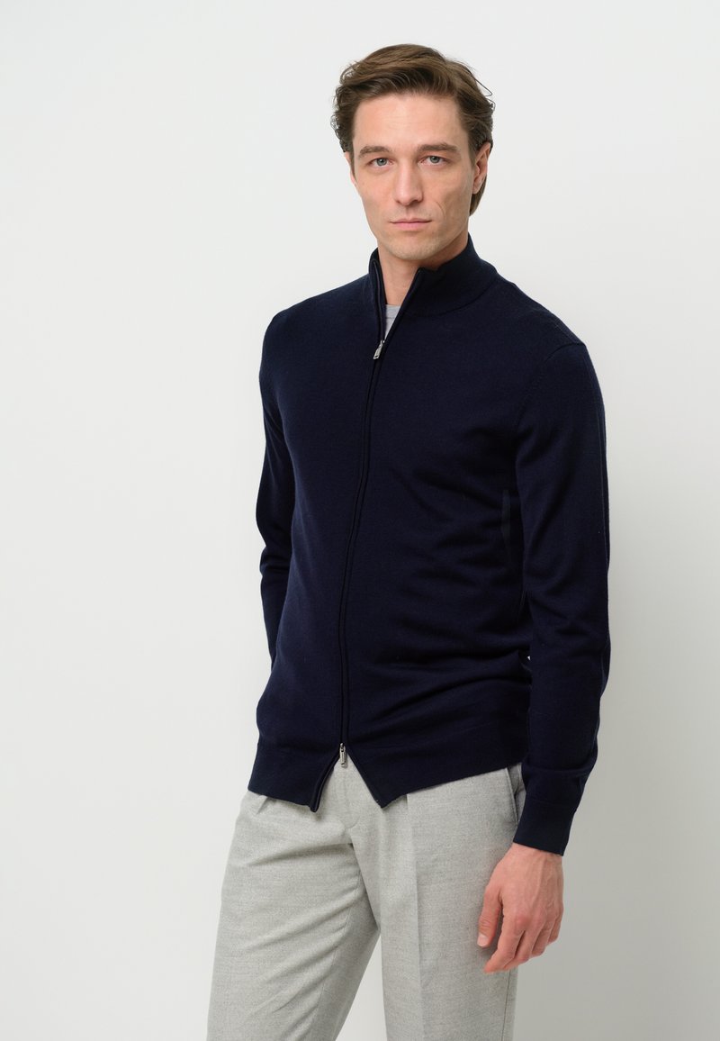 Suéter de cremallera azul marino con cuello alto, puños y dobladillo acanalados. Lleva encima una camisa gris claro, combinada con pantalones gris claro.