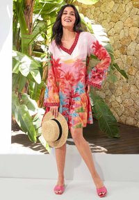 Bunt gekleidetes Kleid mit einem Palmenbaum-Print in den Farben Pink, Rot und Blau. Es hat einen V-Ausschnitt, weit ausgestellte Ärmel und einen plissierten Saum. Ein Strohhut mit einem schwarzen Band ergänzt das Outfit.