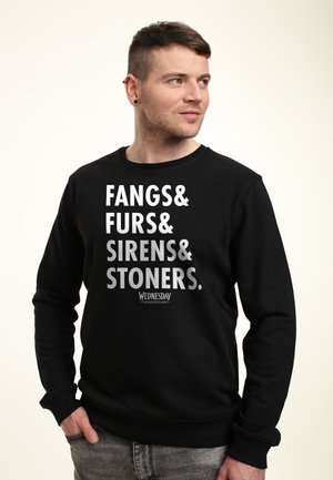 Man met kort haar, die een zwart sweatshirt draagt met de tekst "Fangs & Furs & Sirens & Stoners. Woensdag," kijkt naar rechts met zijn hand in zijn zak.