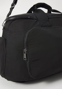 Sac de voyage en nylon noir avec un extérieur rembourré, des compartiments à double fermeture éclair et des poignées courtes. Dispose d'une sangle d'épaule détachable.