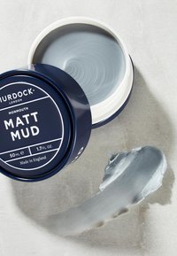 Pot de marin étiqueté "MATT MUD" avec une crème coiffante grise à l'intérieur, à côté d'une tache de crème sur une surface claire. 50 ml, fabriqué en Angleterre.