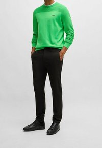 Grön stickad tröja med texturerade ränder på axlarna, svart logotyp. Svarta skräddarsydda byxor och svarta sneakers. Enkel, modern design.