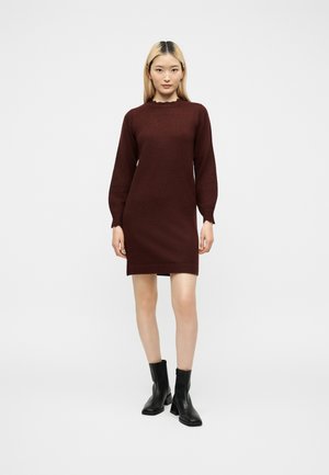 Jeune femme debout, portant une robe en maille bordeaux foncé à manches longues et des bottines noires, sur un fond clair uni.