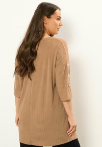 Hellbraunes, geripptes Oberteil mit Drei-Viertel-Ärmeln, das über Knopfakzente an den Ärmeln verfügt. Weicher Stoff und lockere Passform für hohen Tragekomfort.