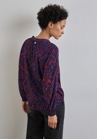 Blusa viola a fantasia con accenti rossi, maniche lunghe e polsini elasticizzati. Collo rotondo con un piccolo dettaglio a bottone sul retro.