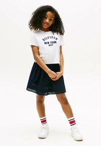 T-shirt bianco a maniche corte con stampa "HILFIGER NEW YORK CITY", abbinato a una gonna blu scuro. Sneakers bianche e calzini a righe completano l'outfit.