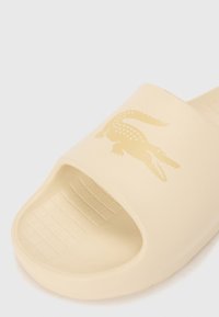 Beige glid sandal tillverkad av gummi, med en strukturerad fotbädd och en gyllene krokodil-logotyp på den övre remmen.