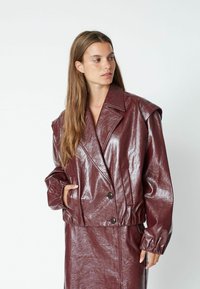 Giacca in ecopelle bordeaux con design doppiopetto, colletto oversize e polsini elastici. Texture liscia e lucida con tasche.