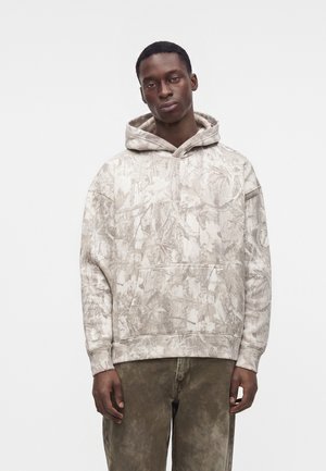 Je jeune homme portant un sweat à capuche gris clair à motif camouflage et un pantalon marron, debout devant un fond blanc uni.