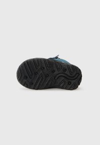 Semelle en caoutchouc noire avec un motif de grip texturé, comportant des indentations circulaires et des lignes ondulées. Partie supérieure lisse et bleue visible le long du bord.