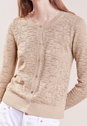 Cardigan en maille beige boutonné avec motif ajouré texturé porté avec un pantalon blanc, torse supérieur de la personne visible.