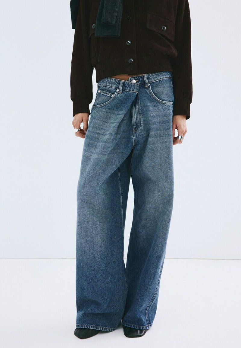 Oseba, oblečena v prevelike modre široke kavbojke z zvitim sprednjim delom, črne čevlje, temno rjav gumb na jopici in prstane na prstih.