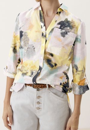 Femme portant une chemise à boutons à imprimé abstrait pastel avec des manches retroussées, rentrée dans un jean gris clair avec une ceinture tressée marron.