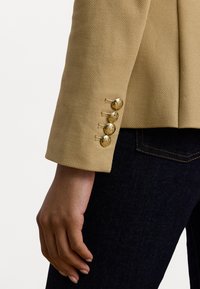 Manche de blazer texturé beige avec quatre boutons dorés et des coutures contrastantes, portée sur un jean foncé. Gros plan de la main reposant sur la cuisse.