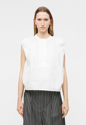 Kvinne med rett lyst brunt hår har på seg en hvit ermeløs bluse og sorte bukser med tynne striper, står foran en ensfarget grå bakgrunn.