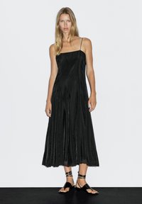 Sort udenærmed kjole med en flydende silhuet, lavet af glat stof. Har tynde stropper og subtile plisseringer for struktur.