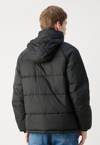 Zwarte pufferjack met capuchon, voorzien van gestikte delen, een gladde textuur en elastische manchetten. De jack wordt gedragen met blauwe jeans.