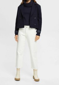 Veste courte croisée bleu marine, col roulé en maille bleu marine, pantalon blanc à jambes droites, et bottines beiges avec semelles marron.