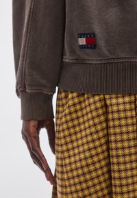 Sudadera marrón con puños y dobladillo acanalados. Presenta un parche con el logo de Tommy Jeans en azul marino, rojo y blanco. Combinada con una falda de patrón texturizado.