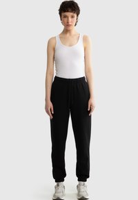 Persoon staand, gekleed in een wit hemdje, zwarte joggingbroek en witte sneakers tegen een effen lichtgrijze achtergrond.