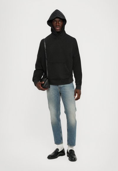 Schwarzer Hoodie mit einer Fronttasche und Kapuze, kombiniert mit hellblauen Jeans, weißen Socken und schwarzen Slippers. Einfaches und modernes Design.