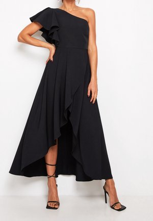 Frau trägt ein schwarzes asymmetrisches One-Shoulder-Kleid mit Rüschensärmeln und schwarzen, hohen Riemchensandalen und steht vor einem weißen Hintergrund.