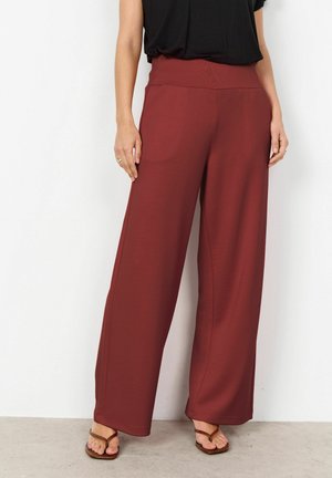 Femme portant un pantalon large taille haute couleur bordeaux associé à un haut noir et des sandales marron, debout devant un fond blanc.