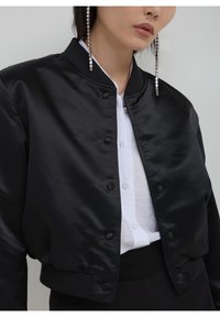 Veste bomber en satin noir au design court, avec un devant boutonné, des poignets côtelés et un col montant.