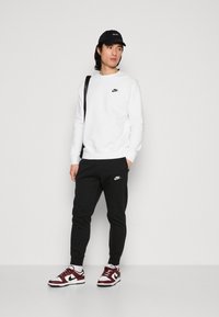 Valkoinen fleece-huppari, jossa on musta Nike-logo, mustat verkkarit, joissa on Nike-logo, ja viininpunaiset-valkoiset lenkkarit. Mallilla on päässään musta lippis.
