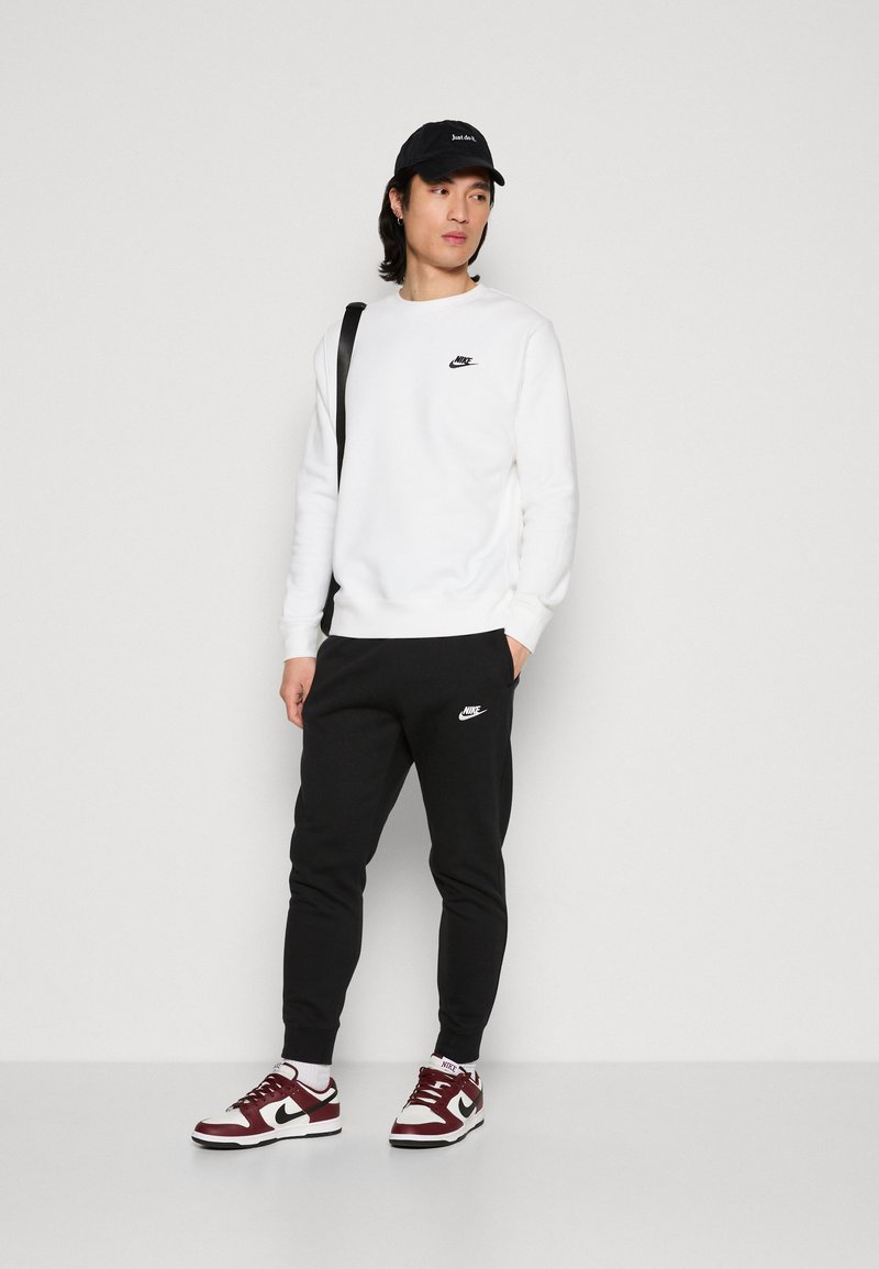 Sudadera blanca de felpa con el logo negro de Nike, pantalones joggers negros con el logo de Nike y zapatillas marón y blancas. El modelo lleva una gorra negra.