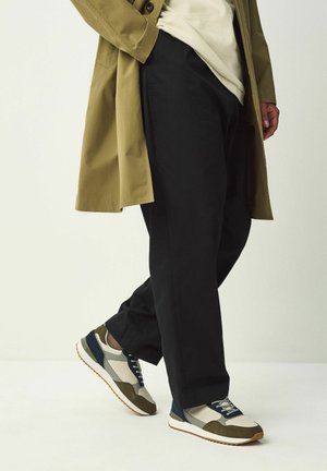 Person iført sorte bukser, beige og olivengrønne sneakers, en beige skjorte og en olivengrøn trenchcoat, stående mod en ensfarvet baggrund.