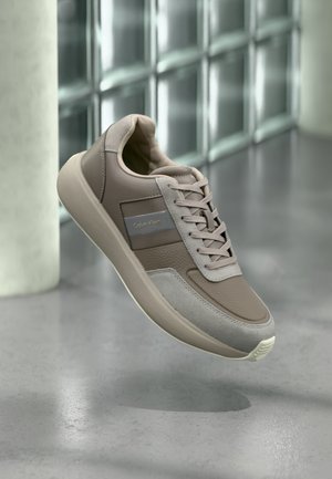 Adidași din piele bej cu o textură netedă și accente din suéd gri. Prezintă o talpă cu amortizare și un design minimalist. Brandul Calvin Klein este vizibil.