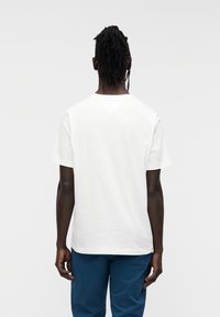 Άσπρο βαμβακερό μπλουζάκι με κοντά μανίκια, άνετη ε φαρμογή και διακριτική V-γραμμή πίσω. Φορεμένο με μπλε παντελόνι, βλέπεται από πίσω.