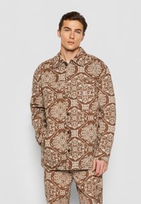 Brun mønstret jakke med indviklede elfenbensdesigns, en kravehals, frontknapper og sidelommer. Glat tekstur og afslappet pasform.