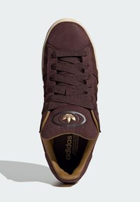 Dark brown sneaker with textured suede material, featuring woven laces, gold logo on tongue, and light beige sole.