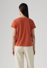 T-shirt en coton rouille à manches courtes avec un col arrondi. Présente une coupe décontractée et des fentes sur les côtés, associé à un pantalon de couleur crème.